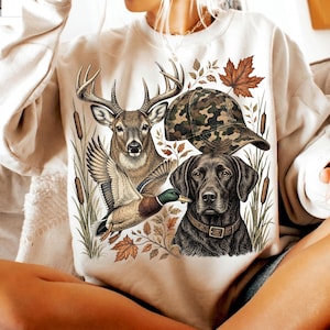 May include: A cream-colored sweatshirt featuring a graphic print of a deer, a mallard duck, a black Labrador retriever wearing a collar, and a camouflage baseball cap, all surrounded by cattails and autumn maple leaves.