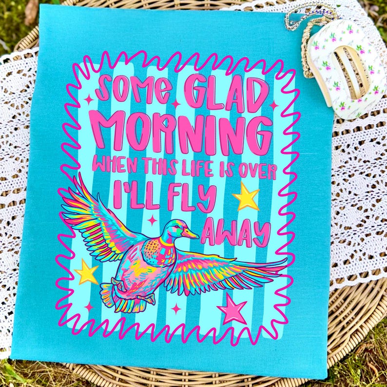 Pu&ograve; includere: Una t-shirt turchese con la grafica di un'anatra in volo dal piumaggio vivace e multicolore. Il testo recita: SOME GLAD MORNING WHEN THIS LIFE IS OVER I'LL FLY AWAY. Il design include stelle rosa e un bordo rosa ondulato.