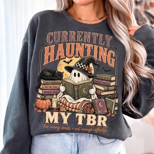Pode incluir: Uma camisola de gola redonda cinzento-carv&atilde;o com uma impress&atilde;o gr&aacute;fica com o texto "CURRENTLY HAUNTING MY TBR" e "too many books - not enough afterlife". O design mostra um fantasma com um chap&eacute;u de bruxa a ler um livro.
