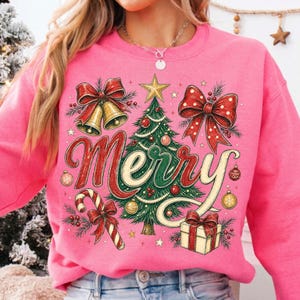 May include: A bright pink sweatshirt featuring a festive graphic print of a green Christmas tree topped with a gold star, surrounded by red bows, gold bells, a candy cane, and a gift box, with the word Merry written in large red script.