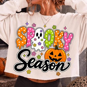 May include: A cream-colored long-sleeve sweatshirt featuring a graphic print with the text "SPOOKY SEASON". The design includes a white ghost, an orange jack-o'-lantern, and colorful polka-dot lettering in orange, pink, and lime green.
