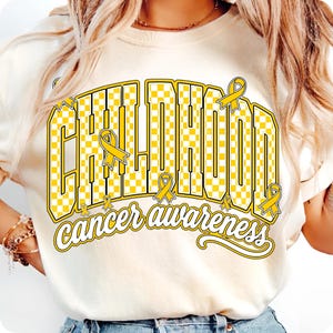 Puede incluir: Una camiseta de color crema con un dise&ntilde;o gr&aacute;fico que presenta la palabra CHILDHOOD en un patr&oacute;n de cuadros amarillos y blancos, acompa&ntilde;ada del texto cancer awareness en letra cursiva, decorada con varios lazos amarillos.