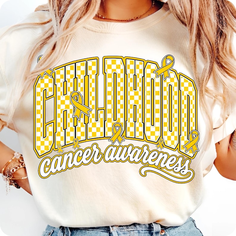 Puede incluir: Una camiseta de color crema con un dise&ntilde;o gr&aacute;fico que presenta la palabra CHILDHOOD en un patr&oacute;n de cuadros amarillos y blancos, acompa&ntilde;ada del texto cancer awareness en letra cursiva, decorada con varios lazos amarillos.