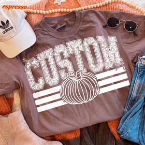 May include: A brown t-shirt featuring the word CUSTOM in large white block letters filled with a repeating pumpkin pattern, accompanied by a large white pumpkin graphic and three horizontal stripes, a beige baseball cap, and sunglasses.