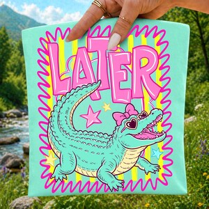 May include: A light mint green tote bag featuring a graphic of a teal alligator wearing heart-shaped sunglasses and a pink bow. The word LATER is printed in large pink letters above the alligator, surrounded by a yellow striped background and pink border.