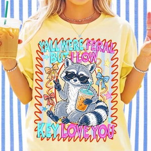 May include: A pale yellow t-shirt featuring a graphic of a raccoon wearing black sunglasses, holding a plastic cup with a straw, and making a peace sign. The text reads YALL WERE FERAL BUT I LOW KEY LOVE YOU, surrounded by colorful bows and stars.