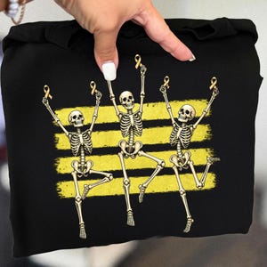 May include: A black sweatshirt featuring a graphic print of three off-white skeletons dancing with their arms raised. Each skeleton holds a small gold awareness ribbon. Behind the skeletons are three horizontal, distressed yellow brush strokes.