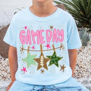 May include: A light blue t-shirt featuring the pink text GAME DAY above a graphic of three stars hanging from a clothesline. The stars include a light green play diagram, a brown football lace design, and a dark green football field pattern.