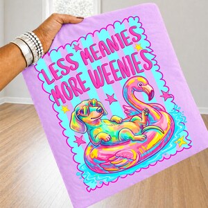 May include: A light purple fabric featuring a graphic of a colorful dachshund wearing heart-shaped sunglasses, lounging on a pink flamingo pool float. The text reads LESS MEANIES MORE WEENIES in bold pink letters, surrounded by pink and yellow stars.