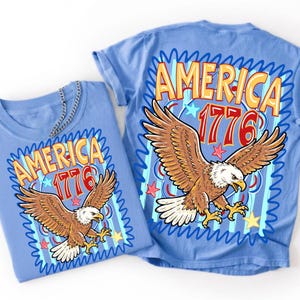 Może przedstawiać: Jasnoniebieski t-shirt z nadrukiem graficznym przedstawiającym lecącego bielika amerykańskiego oraz napis AMERICA 1776 w kolorze ż&oacute;łtym i czerwonym. Wz&oacute;r zawiera niebieskie faliste obw&oacute;dki, czerwone i ż&oacute;łte gwiazdy na jasnoniebieskim tle w paski.