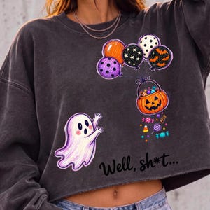 May include: A charcoal gray crewneck sweatshirt featuring a graphic of a white ghost reaching toward a pumpkin-shaped candy bucket held by a cluster of colorful balloons. The text "Well, sh*t..." is printed in black below the ghost and bucket.