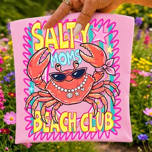 May include: A light pink fabric item featuring a graphic of a red crab wearing black sunglasses and a pearl necklace with a seashell charm. The text reads SALTY MOMS BEACH CLUB in yellow and blue letters, surrounded by pink and teal decorative borders.