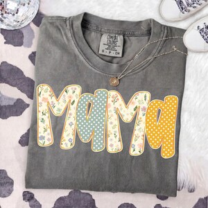 May include: A charcoal gray short-sleeve t-shirt with a crew neckline and a Comfort Colors brand label. The front features the word MAMA in large, rounded letters with alternating patterns of colorful floral prints, light blue stars, and yellow dots.