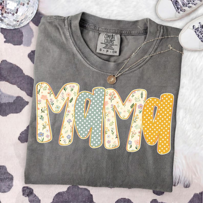 May include: A charcoal gray short-sleeve t-shirt with a crew neckline and a Comfort Colors brand label. The front features the word MAMA in large, rounded letters with alternating patterns of colorful floral prints, light blue stars, and yellow dots.