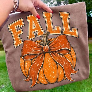May include: A brown t-shirt featuring the word FALL in large, distressed orange block letters arched above a graphic of a bright orange pumpkin adorned with a prominent, matching orange bow tied around its stem in a detailed illustration.
