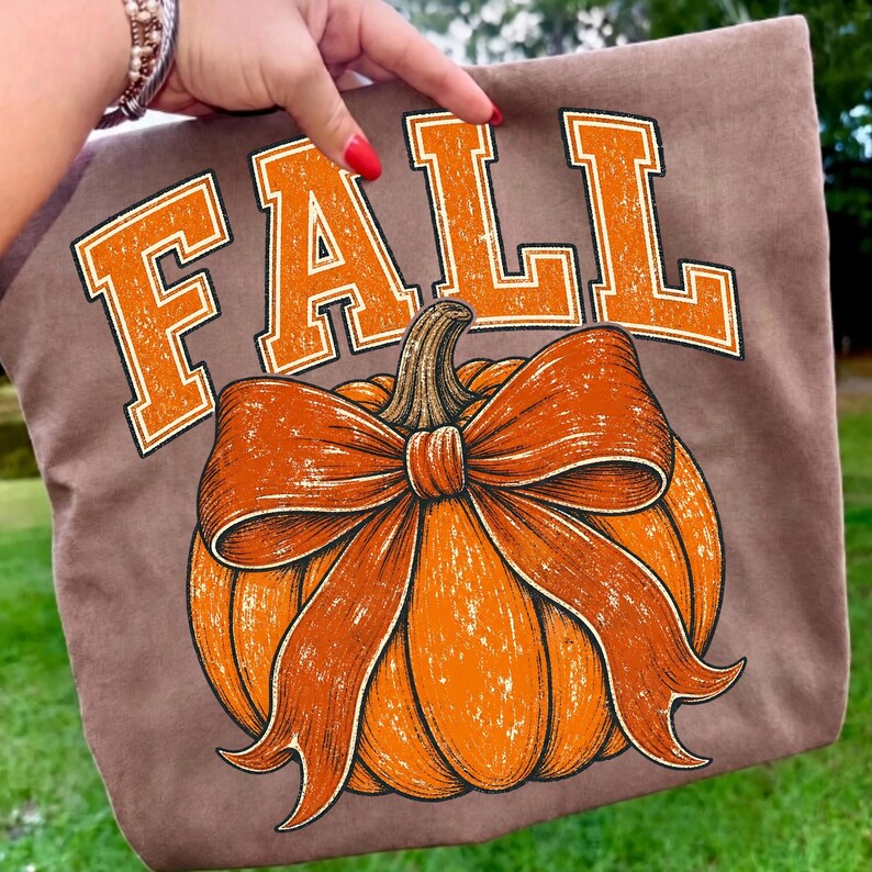 May include: A brown t-shirt featuring the word FALL in large, distressed orange block letters arched above a graphic of a bright orange pumpkin adorned with a prominent, matching orange bow tied around its stem in a detailed illustration.