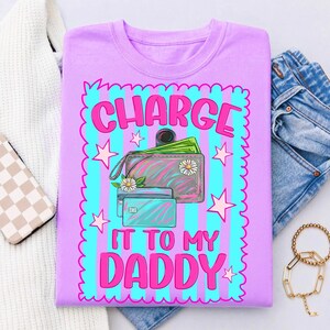 May include: A light purple t-shirt featuring a graphic print with a scalloped border, vertical cyan and purple stripes, and pink stars. The design includes a wallet with green paper money, two credit cards, and the text "CHARGE IT TO MY DADDY".