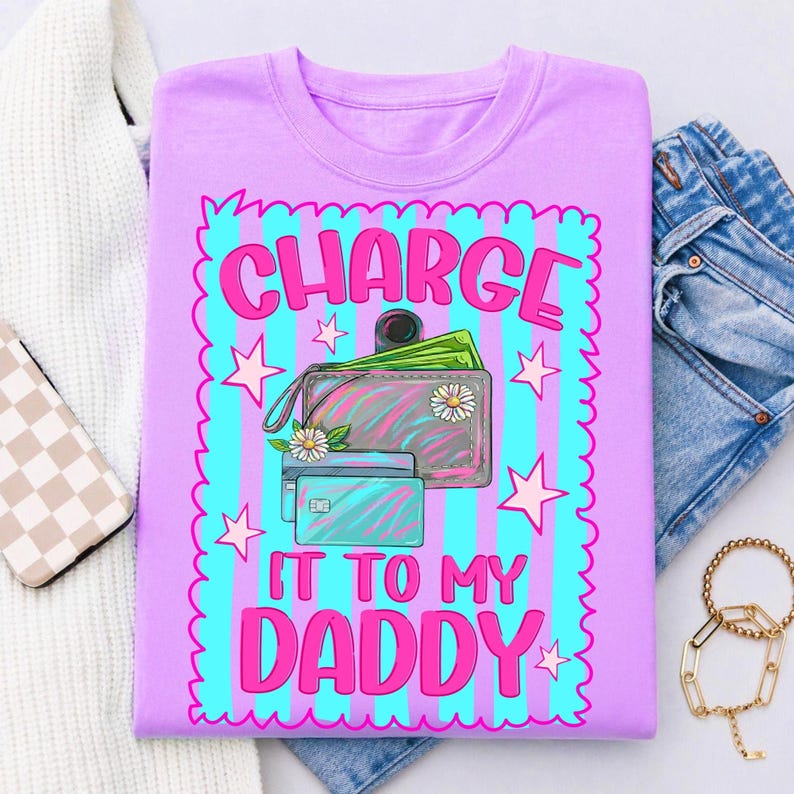 May include: A light purple t-shirt featuring a graphic print with a scalloped border, vertical cyan and purple stripes, and pink stars. The design includes a wallet with green paper money, two credit cards, and the text "CHARGE IT TO MY DADDY".