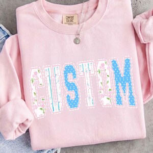 May include: A light pink sweatshirt with the word "CUSTOM" in a decorative font. The letters are a mix of floral and polka dot patterns, outlined with a scalloped edge. A silver necklace with a pendant is visible.