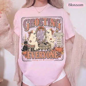 May include: A light pink t-shirt featuring a graphic print with the text GHOSTING EVERYONE. The design shows two ghosts at a vanity mirror, a black cat, pumpkins, and a phone screen graphic displaying 0% battery, 12 calls, 27 texts, and 0 replies.