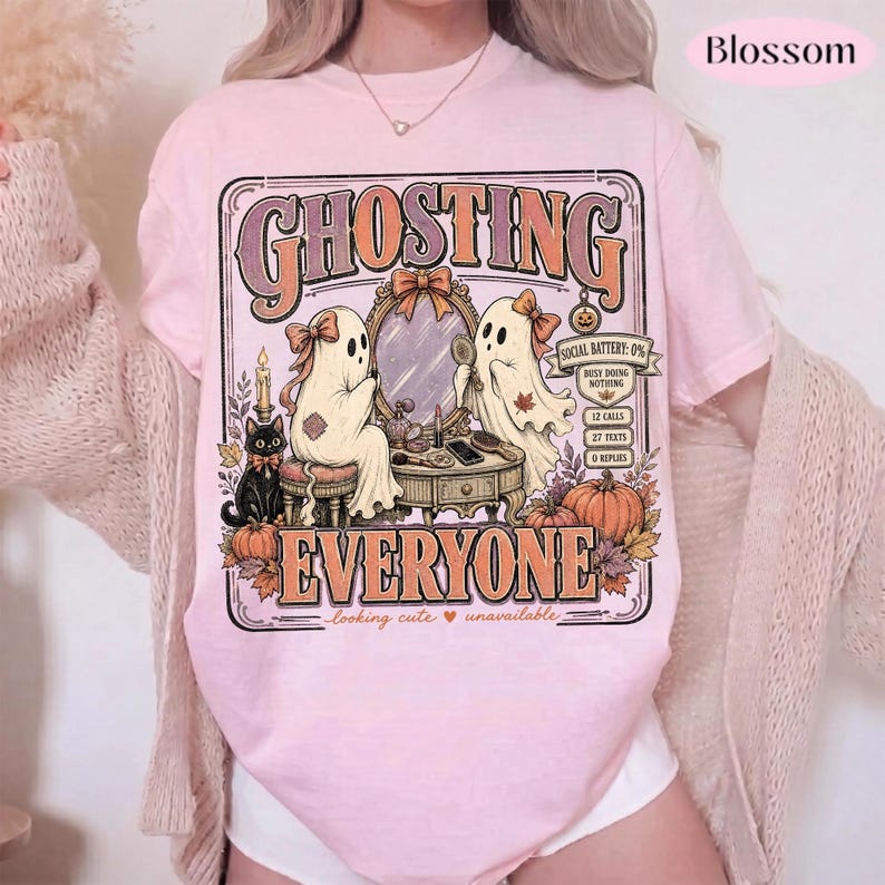May include: A light pink t-shirt featuring a graphic print with the text GHOSTING EVERYONE. The design shows two ghosts at a vanity mirror, a black cat, pumpkins, and a phone screen graphic displaying 0% battery, 12 calls, 27 texts, and 0 replies.