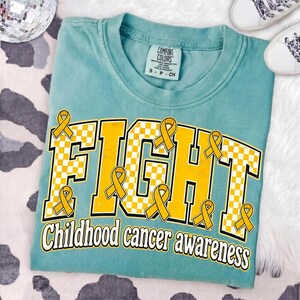 May include: A teal t-shirt featuring the word FIGHT in large yellow and white checkered block letters, with yellow awareness ribbons integrated into the design. Below, the text reads Childhood cancer awareness in a bold yellow font.