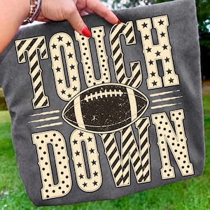 以下が含まれることがあります： クリーム色の大きなブロック体で「TOUCH DOWN」と書かれたダークグレーのTシャツ。文字には星、水玉、斜めストライプの模様が施されています。単語の間には茶色のフットボールのグラフィックが中央に配置されています。