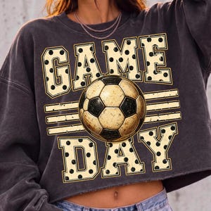 May include: A charcoal gray cropped sweatshirt featuring a graphic print with the text GAME DAY in large, cream-colored block letters with black polka dots. A central soccer ball illustration sits between the words, accented by horizontal lines.