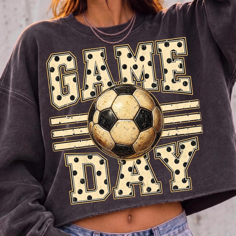 May include: A charcoal gray cropped sweatshirt featuring a graphic print with the text GAME DAY in large, cream-colored block letters with black polka dots. A central soccer ball illustration sits between the words, accented by horizontal lines.