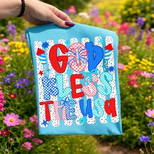 K&ouml;nnte beinhalten: Ein hellblaues T-Shirt mit dem Schriftzug GOD BLESS THE USA in rot-wei&szlig;-blau gemusterten Buchstaben. Das Design zeigt Feuerwerk, Sterne und Raketengrafiken vor einem Hintergrund aus vertikalen schwarz-wei&szlig;en gepunkteten Streifen.