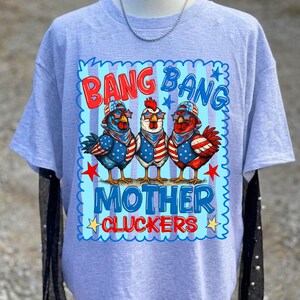 May include: A light gray t-shirt featuring a graphic of three chickens wearing star-spangled bandanas and sunglasses. The text reads BANG BANG MOTHER CLUCKERS in red and blue letters against a light blue striped background with red stars.