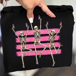 May include: A black sweatshirt featuring a graphic print of three beige skeletons in dynamic poses. Each skeleton holds a small pink ribbon above its head. Behind the skeletons are three horizontal, textured pink brushstroke stripes.