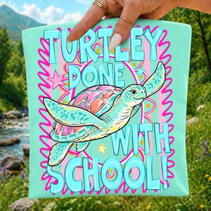 K&ouml;nnte beinhalten: Ein helles mintgr&uuml;nes Stoffquadrat mit einer farbenfrohen Meeresschildkr&ouml;ten-Illustration und dem Text TURTLEY DONE WITH SCHOOL! Das Design enth&auml;lt rosa und gelbe Sternakzente, einen rosa Wellenrand sowie ein zentrales kreisf&ouml;rmiges Muster.