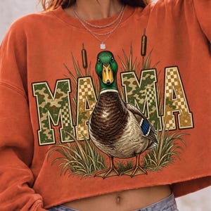 May include: An orange long-sleeve sweatshirt featuring a graphic of a mallard duck standing in tall grass. The word MAMA is printed in large block letters behind the duck, with a camouflage and checkered pattern in shades of green and tan.
