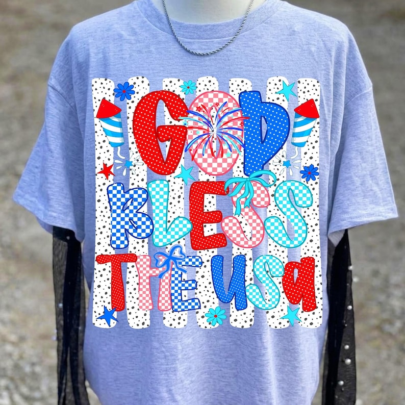 K&ouml;nnte beinhalten: Ein hellgraues T-Shirt mit kurzen &Auml;rmeln und einem Grafikdruck mit dem Text GOD BLESS THE USA. Das Design umfasst rote, blaue und rosafarbene Karomuster, Feuerwerksillustrationen und blaue Schleifen auf einem Hintergrund aus vertikalen schwarzen Punkten.