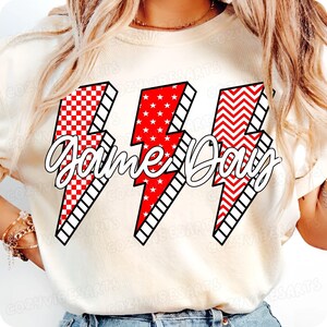 May include: Cream-colored t-shirt featuring three red and white lightning bolt graphics with the text "Game Day" in a cursive font. The lightning bolts have different patterns: checkered, stars, and zigzags.