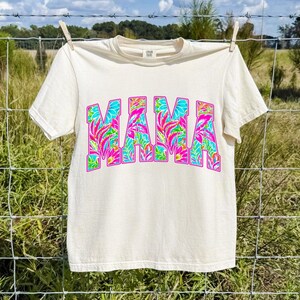 以下が含まれることがあります： クリーム色のTシャツ。大きなカラフルな文字で「MAMA」と書かれています。文字はピンク、青、緑の色合いの熱帯の花柄で埋められています。シャツは屋外の物干しロープに吊るされています。