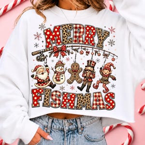 May include: A white long-sleeve sweatshirt featuring a graphic print with the text MERRY FISHMAS. The design includes a fishing rod with ornaments, a Santa Claus, a snowman, a gingerbread man, a nutcracker, and an elf, all in red and leopard print.