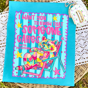 K&ouml;nnte beinhalten: Ein blaugr&uuml;nes T-Shirt mit einer bunten Grafik eines Waschb&auml;ren, der in einem rosa Strandstuhl sitzt und einen gelben Becher h&auml;lt. Der Text lautet: I WANT YOU TO KNOW THAT SOMEONE CARES NOT ME, BUT SOMEONE. Das Design enth&auml;lt gelbe Sterne.