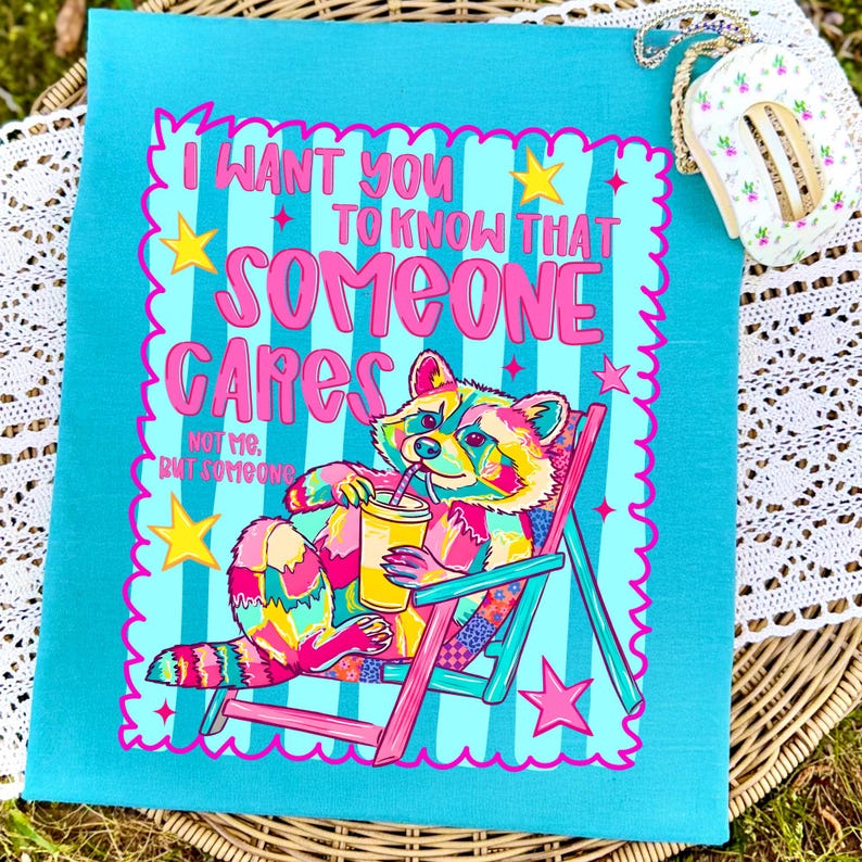 K&ouml;nnte beinhalten: Ein blaugr&uuml;nes T-Shirt mit einer bunten Grafik eines Waschb&auml;ren, der in einem rosa Strandstuhl sitzt und einen gelben Becher h&auml;lt. Der Text lautet: I WANT YOU TO KNOW THAT SOMEONE CARES NOT ME, BUT SOMEONE. Das Design enth&auml;lt gelbe Sterne.
