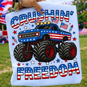 以下が含まれることがあります： アメリカ国旗柄で装飾されたモンスタートラックのグラフィックが描かれたライトブルーのTシャツ。CRUSHIN' FREEDOMという文字が、太字の赤、白、青の配色でプリントされています。デザインには小さなアメリカ国旗と花火が含まれています。