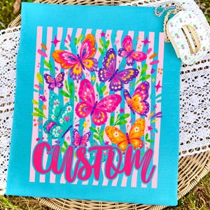 May include: A bright turquoise t-shirt featuring a central graphic of multiple colourful butterflies in shades of pink, purple, orange, and teal against a background of vertical light pink stripes. The word CUSTOM is printed in bold pink script.