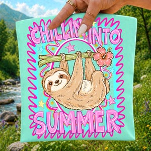 May include: A light mint green tote bag featuring a brown sloth hanging from a branch with a pink hibiscus flower. The graphic includes pink text that reads CHILLIN INTO SUMMER, surrounded by pink squiggly lines and small yellow and pink stars.