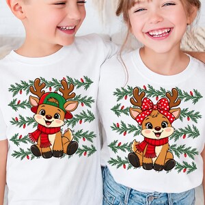 May include: Two white t-shirts featuring a cartoon reindeer graphic. The left shirt shows a reindeer wearing a green baseball cap and red scarf. The right shirt shows a reindeer with a red and white polka dot bow and red scarf. Both have pine branches.