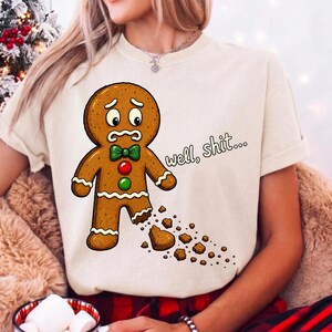 May include: A cream-coloured t-shirt featuring a graphic of a sad gingerbread man with a missing leg and crumbs scattered below. The gingerbread man wears a green bow tie and red and green buttons. The text "well, shit..." is printed in black.