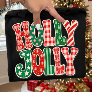 May include: A black sweatshirt featuring the text HOLLY JOLLY in large, colorful, patterned letters. Each letter displays a unique holiday design, including red bows, green polka dots, red gingham, green Christmas trees, and candy cane stripes.