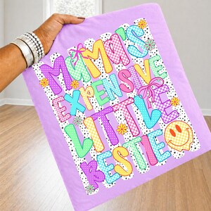 May include: A light purple fabric square featuring a colorful graphic with the text MAMA'S EXPENSIVE LITTLE BESTIE. The design includes checkered patterns in yellow, pink, and blue, small yellow and green flowers, pink bows, and a yellow smiley face.
