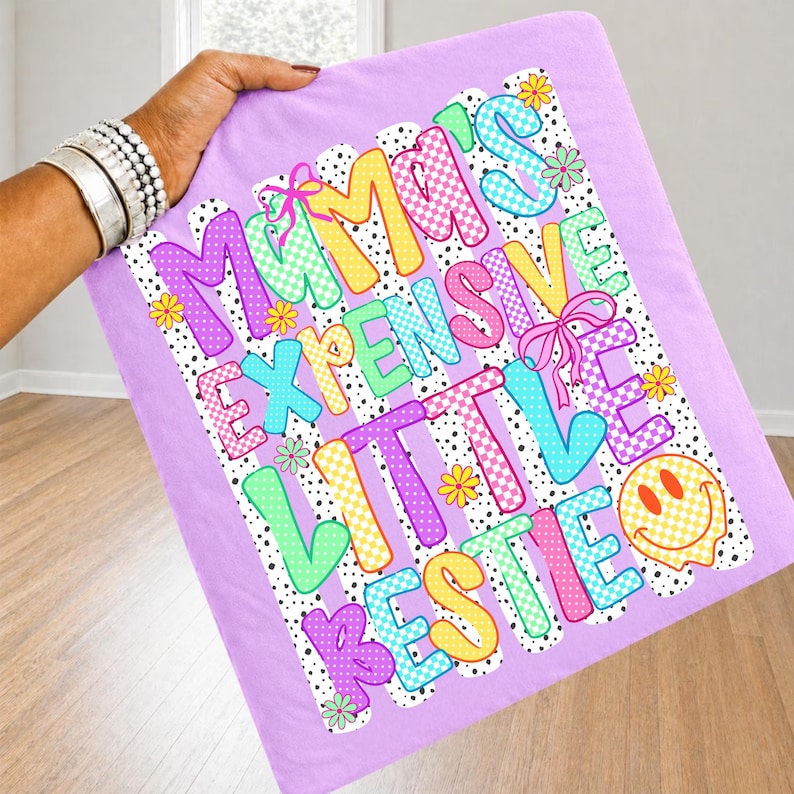 May include: A light purple fabric square featuring a colorful graphic with the text MAMA'S EXPENSIVE LITTLE BESTIE. The design includes checkered patterns in yellow, pink, and blue, small yellow and green flowers, pink bows, and a yellow smiley face.