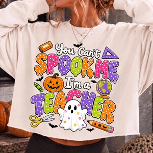 May include: A cream-colored long-sleeve sweatshirt featuring a colorful graphic with the text "You Can't SPOOK ME I'm a TEACHER". The design includes a jack-o'-lantern, a globe, a ghost with flowers, scissors, a pencil, a ruler, and an eraser.