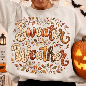 Pu&ograve; includere: Una felpa girocollo grigio chiaro con la scritta Sweater Weather in un carattere decorativo in stile patchwork. Le lettere sono riempite con vari motivi autunnali, inclusi quadri e fiori, circondate da foglie autunnali colorate.
